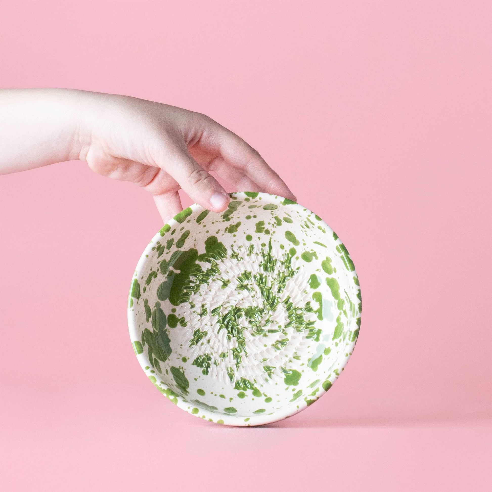 A hand holds the Alfar Tierra Cocida Garlic Grater Plate - Medium Ceramic Green - Moss, featuring handmade Spanish craftsmanship and green splatter patterns, set against a solid pink background.