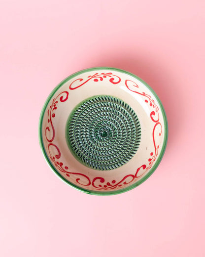 The Alhambra Grater Bowl - Large by Alfar Tierra Cocida is a handcrafted Spanish ceramic piece with an Alhambra-inspired red and green rim, a spiral textured center, and a pastel pink background.