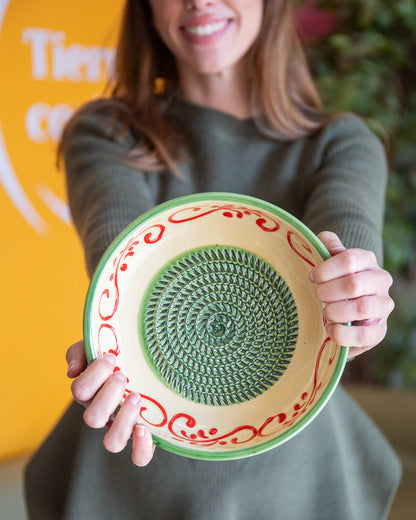 A woman in a green sweater smiles as she holds the Alhambra Grater Bowl - Large by Alfar Tierra Cocida, featuring a green textured center and a cream border with red swirls inspired by the Alhambra’s design.