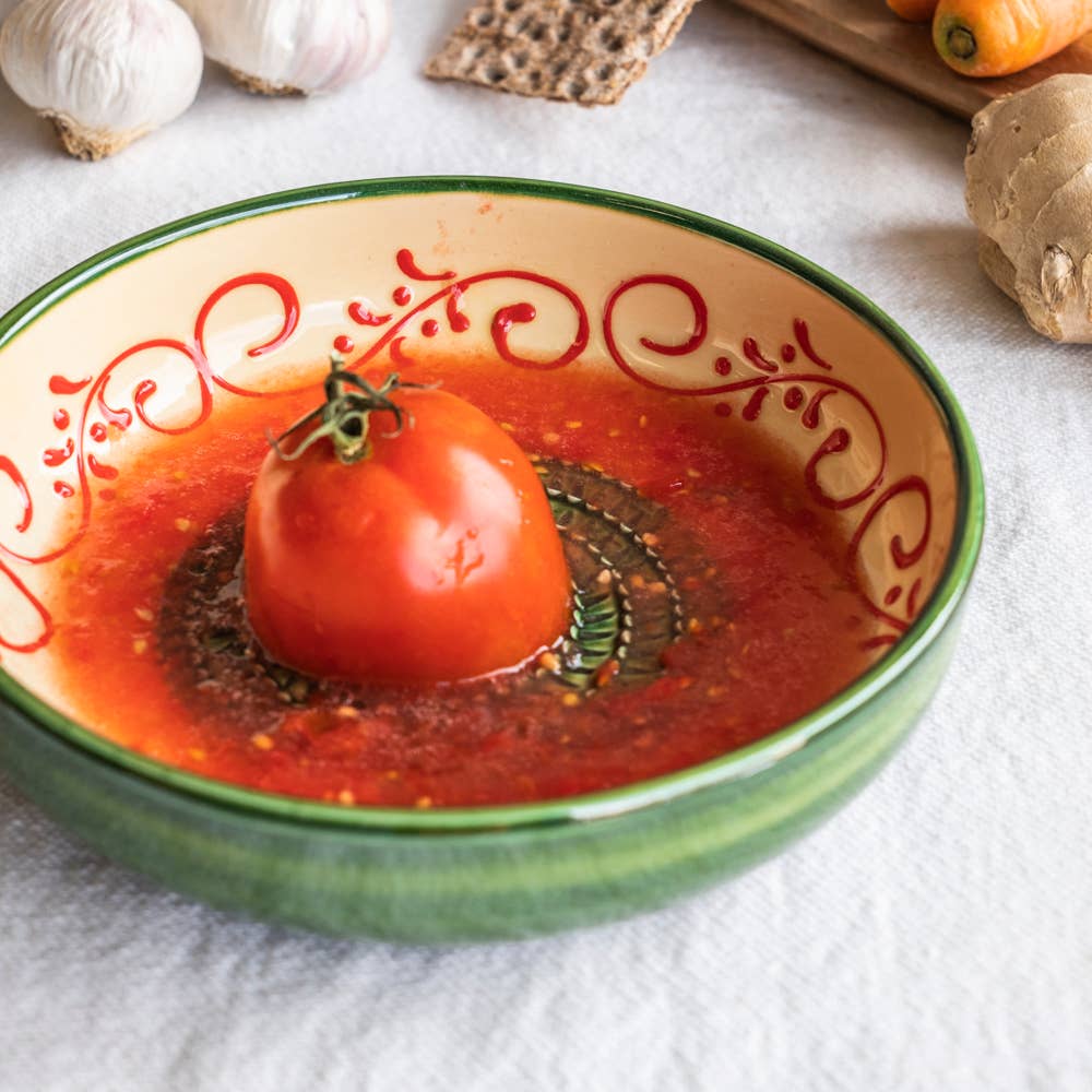 A whole tomato sits in tomato soup, garnished with herbs. The Alhambra Grater Bowl - Large by Alfar Tierra Cocida rests amid garlic, ginger, and vegetables—a tribute to handcrafted Spanish kitchenware.
