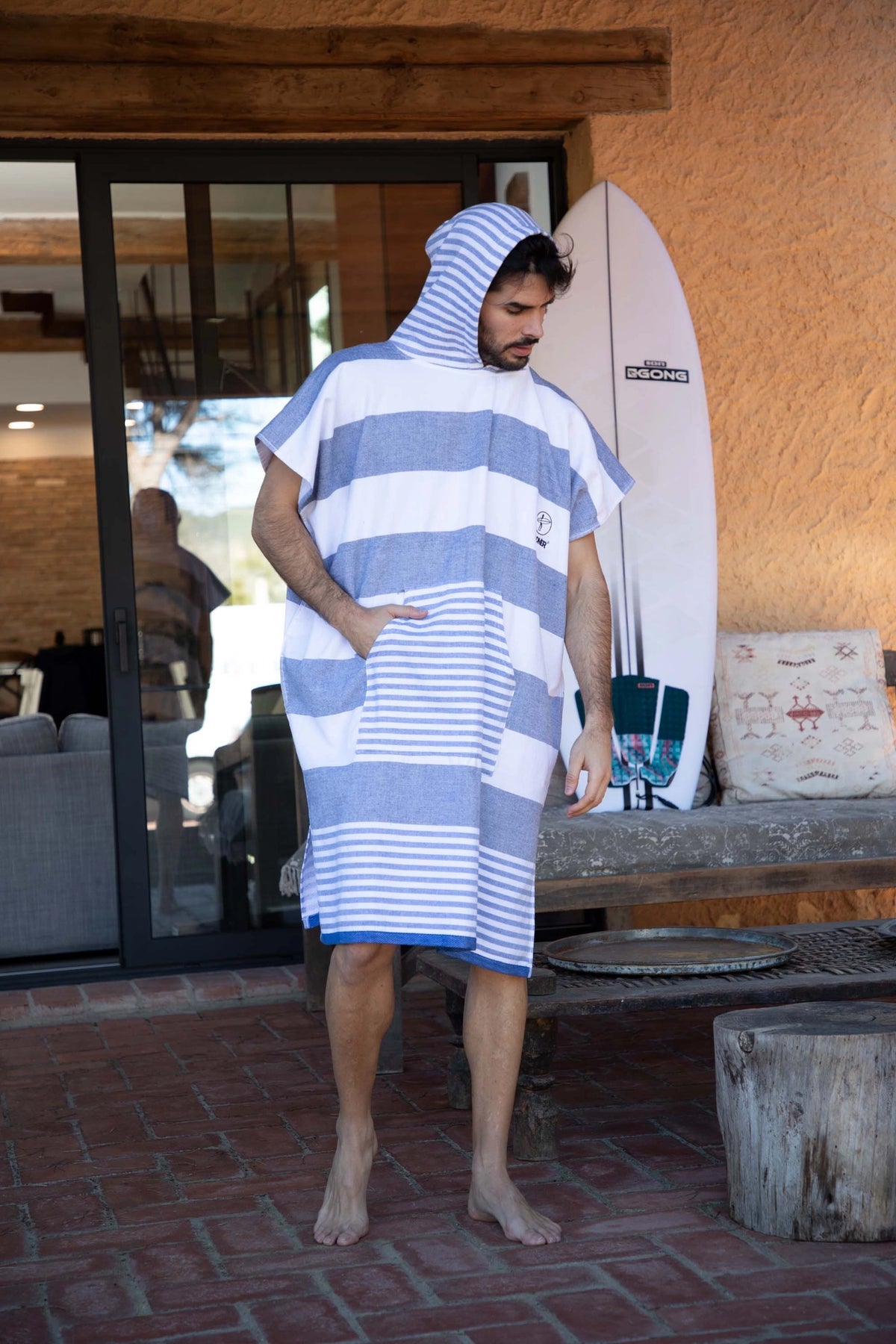 A man wearing a blue and white striped hooded towel poncho standing on a brick patio in front of a building, with a surfboard leaning against the wall behind him.