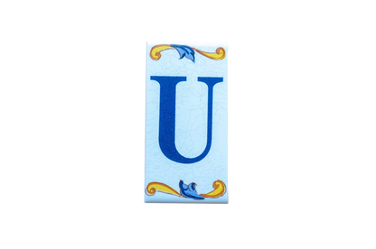 A Lisa Croquets Letters – Ceramic House Letter Tile featuring a blue letter U centered between decorative yellow and blue scrollwork on a light blue background.