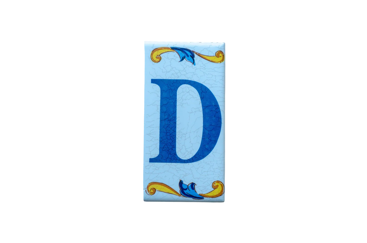 A rectangular Lisa Croquets Letters – Ceramic House Letter Tile featuring the blue letter D centered between decorative yellow and blue scrollwork against a light blue, crackled-texture background.