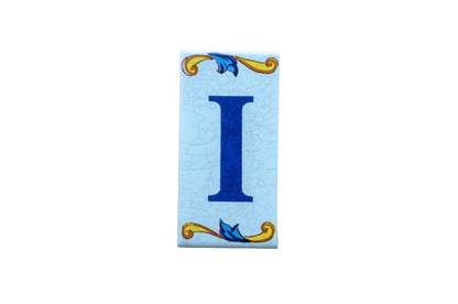 Lisa Croquets Letters – Ceramic House Letter I tile on a light blue crackled background with decorative yellow and blue scrolls at the top and bottom.