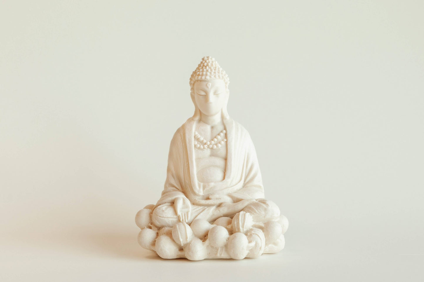 The Manulena Decorative & Aromatic Buddha Piece (White) sits cross-legged on a lotus base with detailed robes and beads, crafted from white ceramic, set against a plain light background.