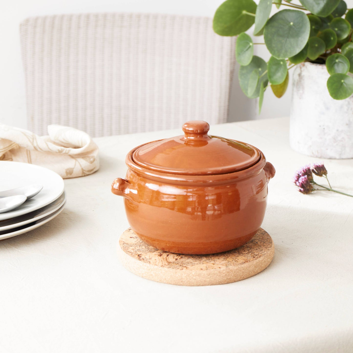 A brown ceramic pot with a lid sits on the LIGA Natural Cork Trivet - Round Small, an eco-sustainable homeware piece, atop a light table beside stacked plates, cutlery, a napkin, a flower, and a potted plant in the background.