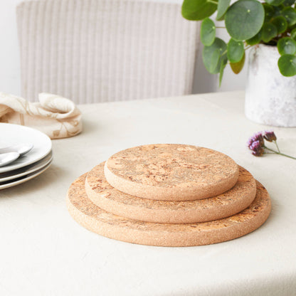 The LIGA Natural Cork Trivet - Round Small offers eco-sustainable, heat-resistant protection. Shown stacked on a light tablecloth, it complements white plates, silverware, a potted plant, and a purple flower for a stylish, green home.