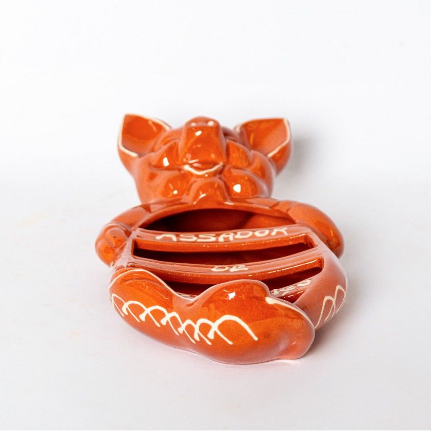 The Ceramica Picas Terracotta Chorizo Pig Roaster is an orange-glazed ceramic tray shaped like a pig with coin slots, white decorative lines, and "ASSADOR" written on its belly.