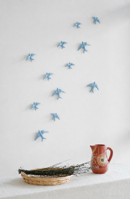 Casa Atlantica’s Ceramic Swallows Individual items - New Colours (Auritas) arranged on a white wall evoke Mediterranean charm, like birds in flight above a table with a woven twig basket and a red spiral ceramic pitcher.
