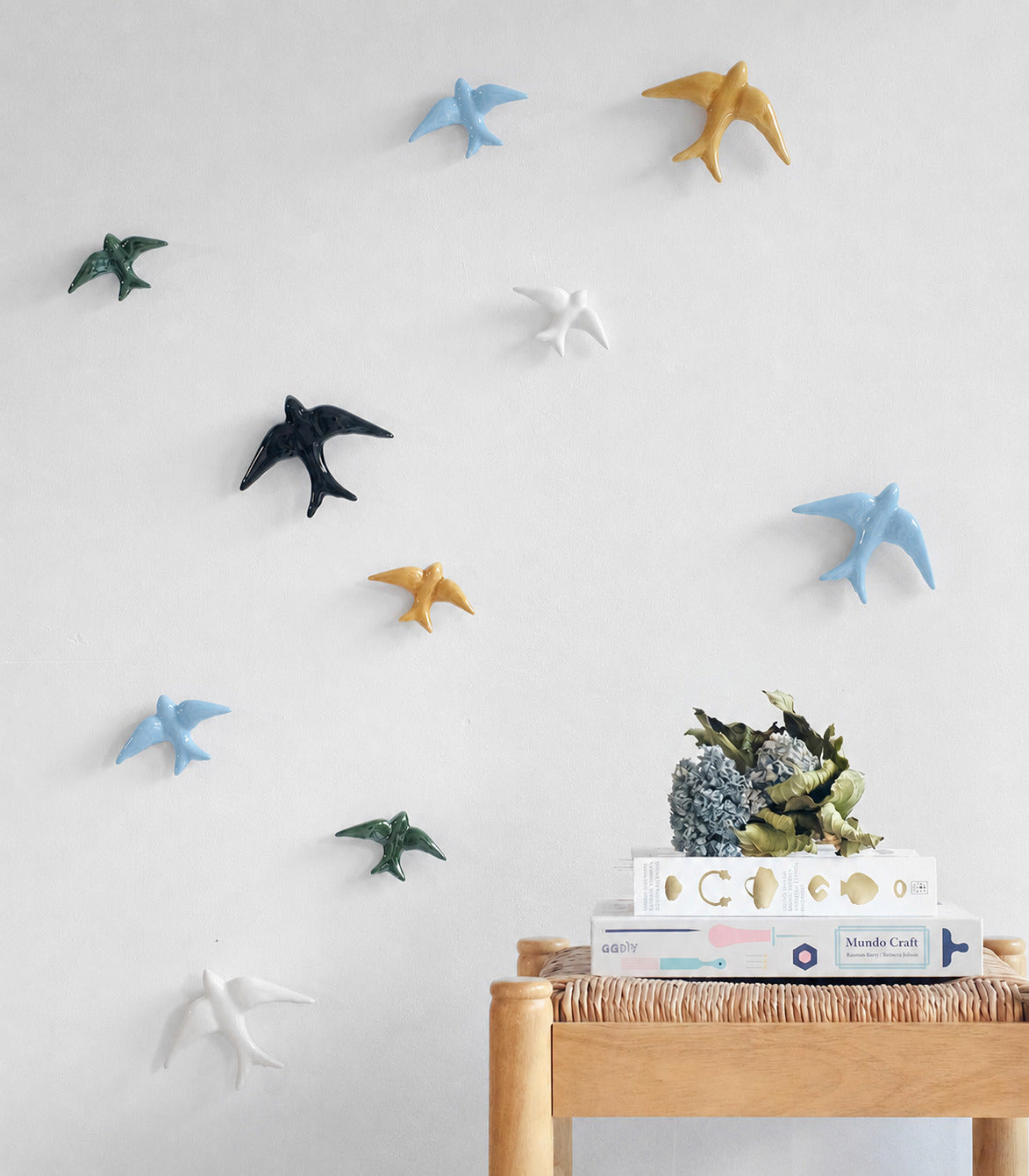 Casa Atlantica’s Ceramic Swallows Individual items – New Colours (Auritas) bring Mediterranean charm to a white wall. Beneath them, a small wooden table displays books and florals, creating a warm and inviting decor.