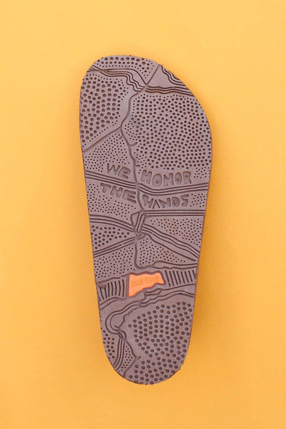 A brown Tampico sandal sole by Mexas EU, featuring textured patterns and "WE HONOR THE HANDS" carved on it, is displayed against a mustard yellow background, highlighting its tribute to handcrafted tradition.